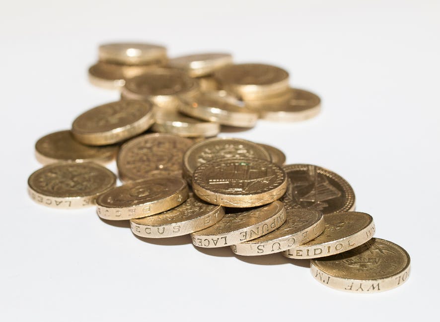 A close-up image of a scattered pile of various UK one-pound coins made of gold-coloured metal, some stacked and others lying flat on a plain white surface. The coins display embossed designs and insc