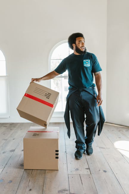 A man with dark curly hair and a beard, dressed in a blue T-shirt and dark trousers, is inside a well-lit room with white walls and large arched windows, preparing to lift a stack of cardboard boxes f